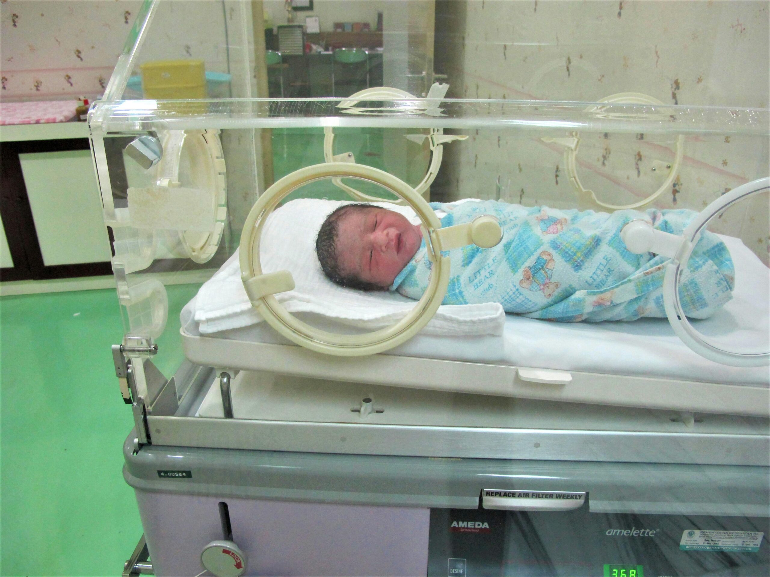 A newborn baby lies in an incubator at a hospital, wrapped in a colorful blanket.